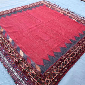 May include: A large, rectangular rug with a vibrant red center and a decorative border. The border features intricate geometric patterns in shades of brown, black, and cream. The rug has a dark blue zig-zag design around the red center.