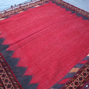May include: A red rug with a dark blue zigzag border and a detailed patterned edge. The rug has a soft, textured appearance and is likely made of wool or a similar material. The design is traditional.
