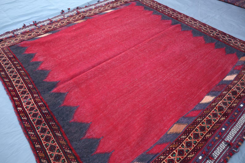 May include: A red rug with a dark blue zigzag border and a detailed patterned edge. The rug has a soft, textured appearance and is likely made of wool or a similar material. The design is traditional.