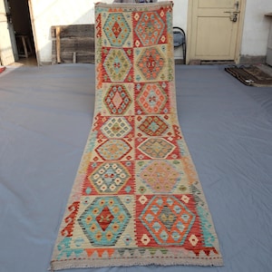 Könnte beinhalten: Ein langer, rechteckiger Teppich mit geometrischem Muster. Der Teppich weist ein sich wiederholendes Design aus farbigen Formen auf, darunter Sechsecke und Rauten, in Rot-, Blau-, Orange- und Beigetönen. Der Teppich liegt auf einer grauen Oberfläche.