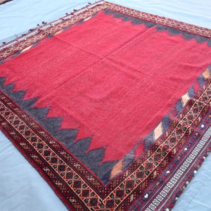 May include: A rectangular, red rug with a dark gray zigzag border and intricate patterned trim. The rug has a central red field and is likely hand-woven, showcasing traditional craftsmanship. The trim features geometric designs in various colors.