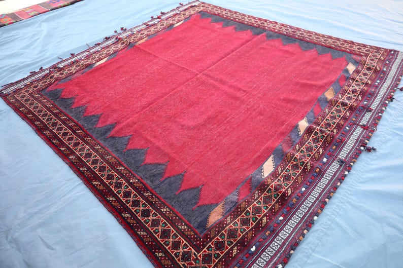 May include: A rectangular, red rug with a dark gray zigzag border and intricate patterned trim. The rug has a central red field and is likely hand-woven, showcasing traditional craftsmanship. The trim features geometric designs in various colors.