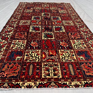 May include: A rectangular, hand-woven rug with a repeating geometric pattern. The rug features a central grid of squares in shades of red, navy, and cream, with floral and abstract designs. The border is a darker red with intricate patterns.