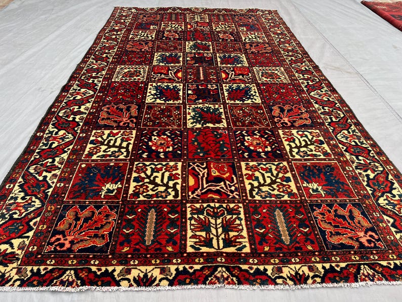 May include: A rectangular, hand-woven rug with a repeating geometric pattern. The rug features a central grid of squares in shades of red, navy, and cream, with floral and abstract designs. The border is a darker red with intricate patterns.