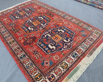 Puede incluir: Una alfombra rectangular, tejida a mano, con fondo rojo y complejos diseños geométricos en azul marino, blanco, amarillo y otros colores. La alfombra presenta un diseño de medallón central y un borde detallado con flecos.