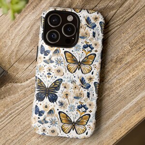 Puede incluir: Una funda de teléfono blanca con un patrón repetido de mariposas doradas, azul marino y blancas, intercaladas con pequeñas flores azules. La funda tiene un acabado brillante y un recorte para la cámara.