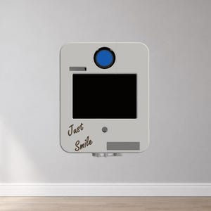 May include: A light gray rectangular photo booth enclosure featuring a prominent blue circular lens at the top, a large black rectangular screen in the center, and the text Just Smile printed in a dark script font on the lower left side.