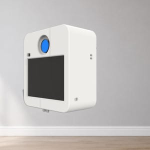 May include: A white, box-shaped photo booth enclosure featuring a central circular lens opening with a blue interior and a large rectangular black screen on the front face. The unit has rounded corners and a smooth, matte white exterior finish.