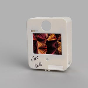 May include: A cream-coloured, rectangular device featuring a screen with a vibrant, abstract design. The words "Just Smile" are written in cursive. There is a circular aperture at the top of the device.