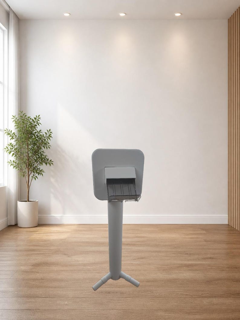 May include: A grey information stand with a rectangular top and a three-legged base. The stand is in a room with a wooden floor, a white wall, and a potted plant. The stand is made of metal.
