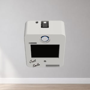 Puede incluir: Un dispositivo blanco y rectangular con una pantalla negra y las palabras "Just Smile" en cursiva. Tiene una ranura, un bot&oacute;n y una peque&ntilde;a abertura en la parte superior. El dispositivo est&aacute; montado en una pared blanca.