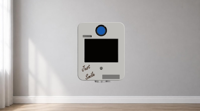 Puede incluir: Un fotomat&oacute;n gris claro con una pantalla negra, un bot&oacute;n azul y las palabras "Just Smile" en cursiva. El fotomat&oacute;n est&aacute; montado en una pared gris claro, con un rodapi&eacute; blanco y una ventana a la izquierda.