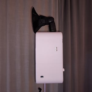 May include: A white photo booth with a black flash and a black shade attached to the top. The photo booth has two silver buttons and a USB port. The photo booth is on a stand in front of a gray curtain.