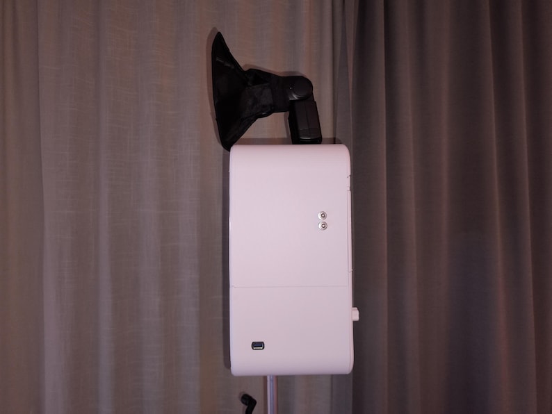May include: A white photo booth with a black flash and a black shade attached to the top. The photo booth has two silver buttons and a USB port. The photo booth is on a stand in front of a gray curtain.