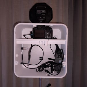 May include: A white, rectangular photo booth with a camera, cables, and a flash unit. The camera is on a shelf inside the booth. The flash unit is black and sits on top of the booth. Cables and wires are visible.