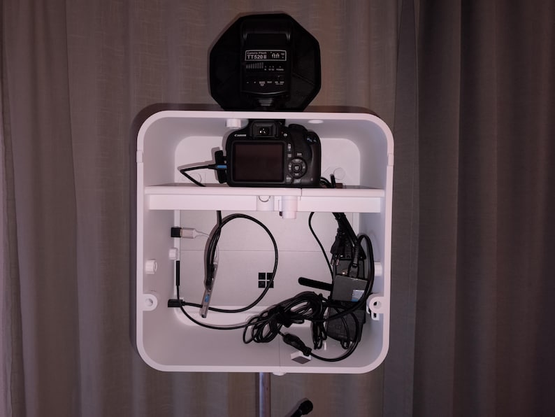 May include: A white, rectangular photo booth with a camera, cables, and a flash unit. The camera is on a shelf inside the booth. The flash unit is black and sits on top of the booth. Cables and wires are visible.