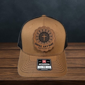May include: A brown trucker hat with a black mesh back. The hat features a leather patch with a sunflower design and a cross, along with the text "SHINE ANYWAY Matthew 5:16". The hat has an adjustable strap.