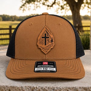 May include: A tan trucker hat with a black mesh back and a brown leather patch on the front. The patch features a mountain landscape, a cross, a pine tree, and the text Psalm 121:1. A black sticker on the brim reads Richardson Original 112 Trucker.