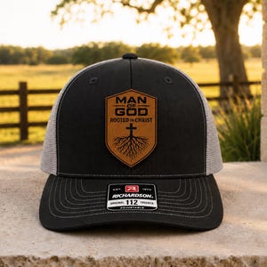 May include: A black trucker hat with a grey mesh back and a brown leather-like shield patch on the front. The patch features the text MAN OF GOD ROOTED IN CHRIST above a cross with tree roots. A black sticker on the brim reads RICHARDSON 112.