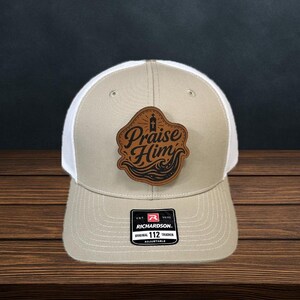 May include: Tan and white trucker hat with a leather patch. The patch features the words "Praise Him" in a stylized font, with a cross and wave design. The hat has a curved brim and an adjustable strap.