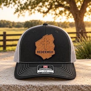 May include: A black and grey trucker hat featuring a tan leather patch on the front. The patch is shaped like a state outline and displays a cross symbol with the text REDEEMED. A black sticker on the brim reads RICHARDSON 112 ORIGINAL TRUCKER.