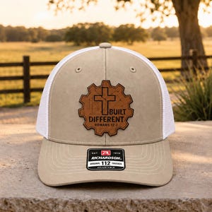 May include: A tan and white trucker hat featuring a brown leather-like patch in the shape of a gear. The patch displays a cross and the text BUILT DIFFERENT ROMANS 12:2. A black sticker on the brim reads RICHARDSON 112 TRUCKER ADJUSTABLE.