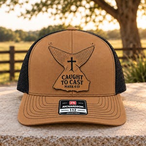 May include: A tan and black trucker hat featuring a leather patch with a fish tail graphic, a cross, and the text CAUGHT TO CAST MARK 4:19. The brim has a black sticker with the text RICHARDSON ORIGINAL 112 TRUCKER ADJUSTABLE.