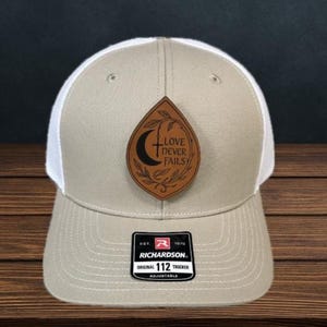 May include: Tan and white trucker hat with a leather patch. The patch features the text "LOVE NEVER FAILS" with a crescent moon and cross design. The hat has a curved brim and an adjustable strap.