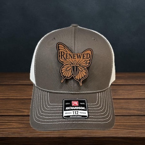 May include: A gray and white trucker hat with a leather butterfly patch that reads "Renewed." The hat has a curved brim and an adjustable strap. The patch is brown with the word "Renewed" in a dark brown font.