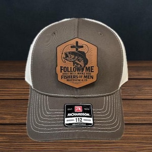 May include: A gray and white trucker hat with a leather patch featuring a fish, cross, and the text "FOLLOW ME AND I WILL MAKE YOU FISHERS OF MEN MATTHEW 4:19." The hat has a curved brim and an adjustable strap.