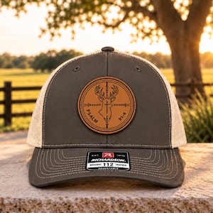 May include: A charcoal gray and beige mesh trucker hat featuring a circular brown leather patch on the front. The patch displays a deer head silhouette inside a crosshair graphic with the text Psalm 91:4. A black sticker is on the brim.
