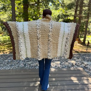 May include: A chunky knit blanket in shades of beige, cream, and brown. The blanket features vertical stripes and a thick, textured weave. The edges are finished with a dark brown border. The person is wearing blue jeans and white shoes.