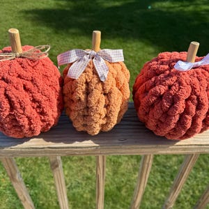 Op de afbeelding: Drie handgemaakte, pluche pompoenen in tinten oranje en rood staan op een houten reling. Elke pompoen heeft een houten steel en is versierd met een decoratieve strik. De pompoenen zijn een gezellige herfstdecoratie.
