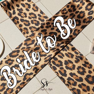 May include: A leopard print sash with the words "Bride to Be" in white script. The sash is brown and black with a repeating animal print pattern. A perfume bottle and a small plate with a gold rim are in the background.