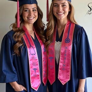 May include: Two women wearing navy blue graduation gowns and caps with pink nursing sashes. The sashes have the word "NURSING" in blue, along with medical symbols and text. The image is from Sash & Style.