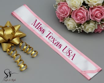 May include: A white sash with pink trim and the text "Miss Texas USA" in a magenta color. A gold bow and gold ringlets are on the left, and a bouquet of pink and white roses is on the right.