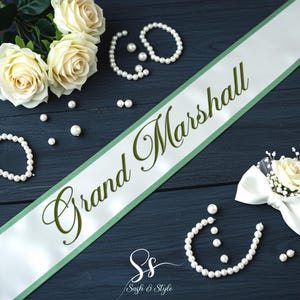 May include: A white sash with "Grand Marshall" in green script, surrounded by pearl bracelets, necklaces, and scattered pearls. A white rose boutonniere and a bouquet of cream roses complete the arrangement. The items are arranged on a dark blue surface.