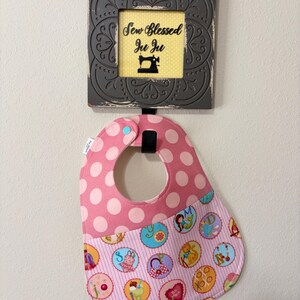 May include: A pink polka dot baby bib with a colorful patterned bottom section featuring circular designs. Above the bib is a framed sign with the text "Sew Blessed Ju Ju" and a sewing machine illustration.