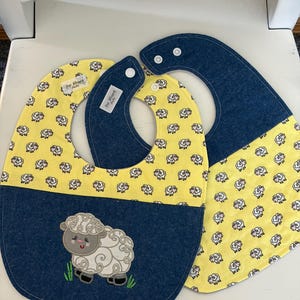May include: Two baby bibs. One bib features a yellow fabric with a sheep pattern and a navy blue bottom with an embroidered sheep. The other bib is yellow with a sheep pattern and navy blue neckband. The bibs have the text "See Blessed Kids".