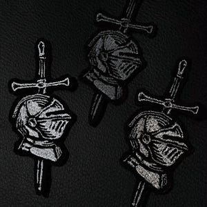 Puede incluir: Tres parches bordados con un diseño de casco de caballero y espada. Los parches son negros con costuras blancas, sobre un fondo negro. Estos parches son probablemente para ropa o accesorios.