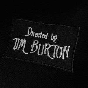 Op de afbeelding: Zwart rechthoekig embleem met witte geborduurde tekst: "Directed by Tim Burton". Het embleem is genaaid op een zwarte, getextureerde stof. De tekst is in een gestileerd lettertype.