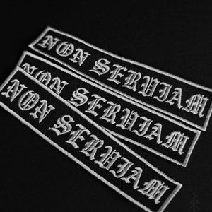 Op de afbeelding: Drie rechthoekige zwarte stoffen patches met de wit geborduurde tekst NON SERVIAM in een gotisch lettertype. Elke patch heeft een wit gestikte rand en is diagonaal gerangschikt op een effen zwarte achtergrond.