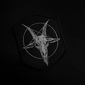 Può includere: Toppa nera con un'immagine ricamata in bianco di un teschio di capra all'interno di un pentagramma. La toppa ha la forma di uno scudo ed è posta su uno sfondo nero. Il design è dettagliato e in stile gotico.