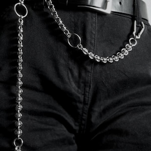 Può includere: Un accessorio a catena in metallo color argento con complessi maglie intrecciate, anelli circolari e una chiusura a moschettone. La catena è fissata a una cintura in pelle nera con fibbia argentata, indossata sopra pantaloni in denim nero.