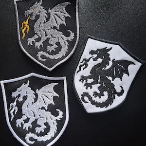 Può includere: Tre toppe ricamate a forma di scudo con disegni di draghi. Una toppa presenta un drago grigio su sfondo nero con fiamme gialle. Un'altra mostra un drago bianco su sfondo nero. La terza raffigura un drago nero su uno sfondo di colore bianco.