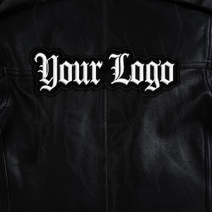 Op de afbeelding: Zwarte leren jas met een wit geborduurd embleem. Het embleem toont de woorden "Your Logo" in een gotisch lettertype. De jas heeft een getextureerde uitstraling en een klassieke biker-stijl.