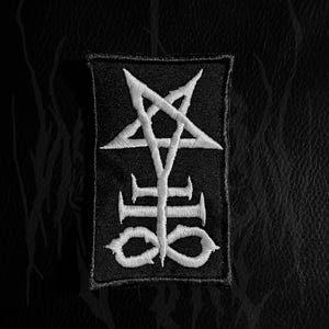 Może przedstawiać: Czarna i biała naszywka z haftowanym, stylizowanym symbolem. Projekt zawiera odwrócony pentagram nad pionową linią z trzema poziomymi paskami i stylizowanym dołem. Naszywka jest prostokątna z czarną obwódką.