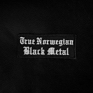 Op de afbeelding: Zwarte rechthoekige patch met witte geborduurde tekst. De tekst luidt "True Norwegian Black Metal". De patch is geplaatst tegen een zwarte achtergrond met textuur, waarschijnlijk leer of een vergelijkbaar materiaal.