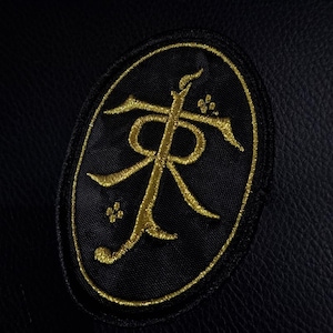 Può includere: Toppa nera con design ricamato in oro. Il design ha una forma ovale con una lettera "R" stilizzata e altri simboli. La toppa è cucita su un materiale nero simile alla pelle. La toppa è probabilmente un elemento decorativo.