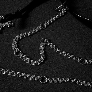 Op de afbeelding: Een zilverkleurige ketting met een reeks kleine, cirkelvormige schakels. De ketting wordt gepresenteerd op een donker, getextureerd oppervlak, met een deel van een zwart object op de achtergrond. De ketting is ontworpen met verschillende cirkelvormige ringen.